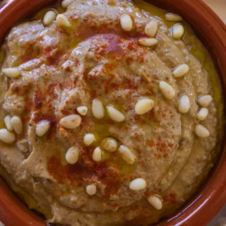 A bowl of baba ganoush is garnished with some olive oil and toasted pine nuts