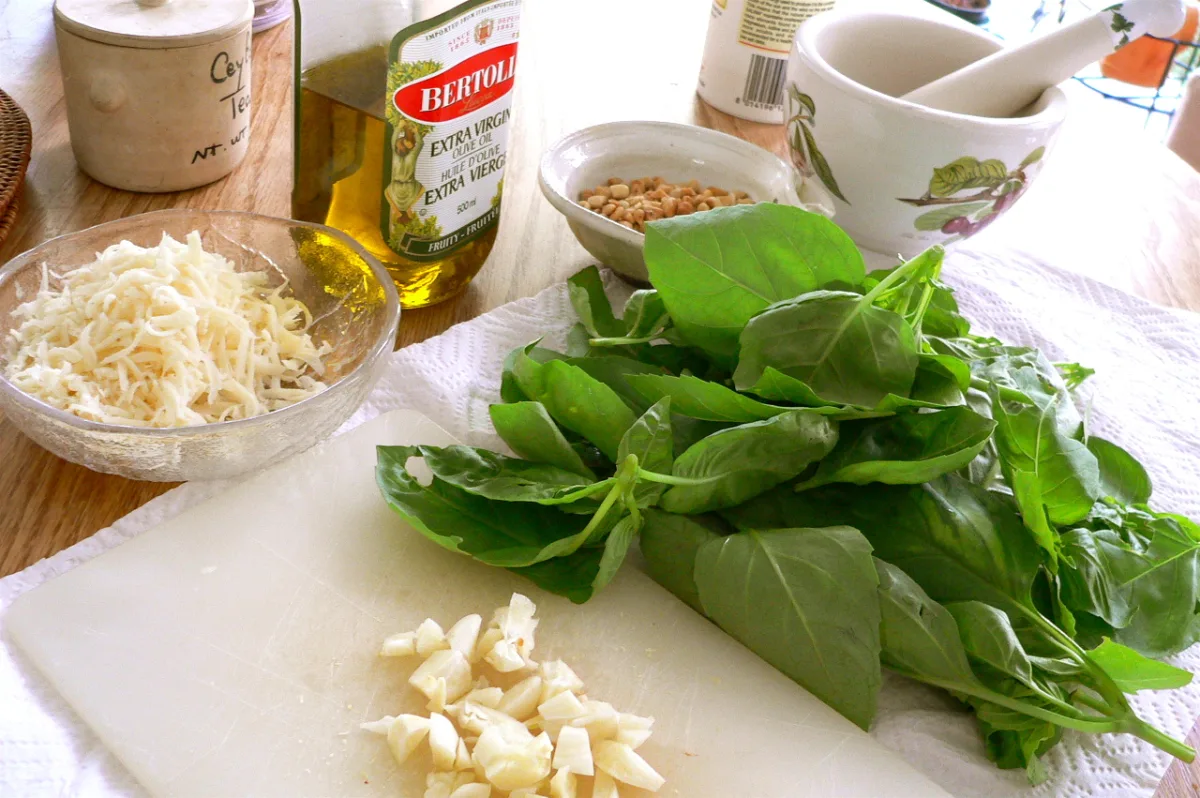 Fresh basil leaves, chopped garlic, parmesan cheese, and a bottle of olivee oil on a kitchen counter.