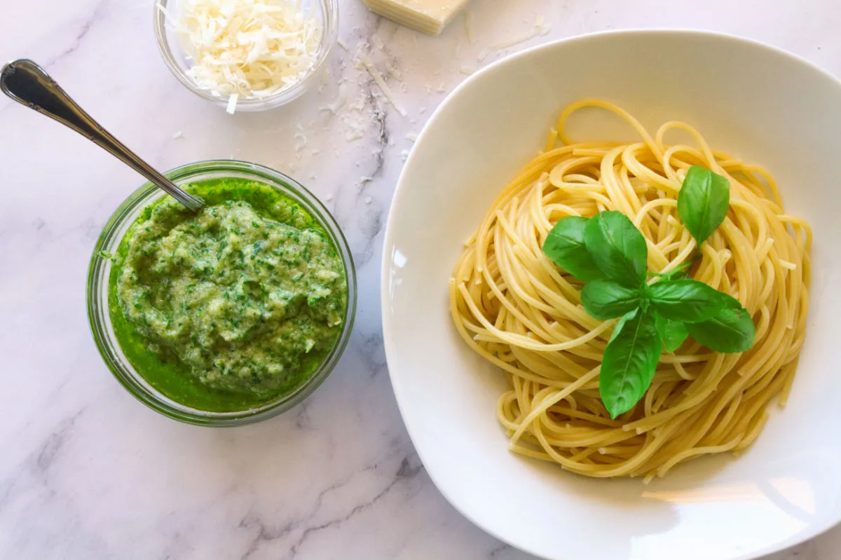 A small bowl of homemade basil pesto sits beside a bowl of spaghetti.