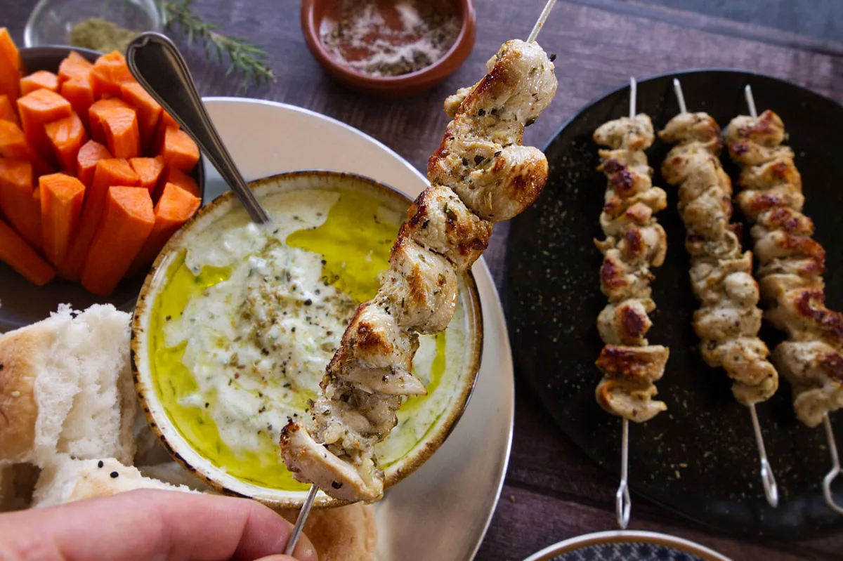 Some chicken souvlaki skewers being served with some tzatziki and some carrot sticks.