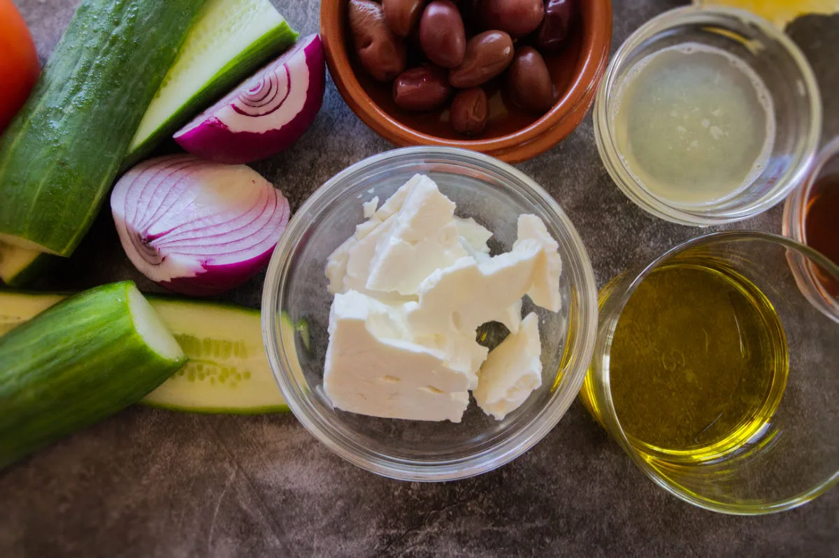 Feta cheese, olive oil, black olives, cucumber, and red onion sit on a counter top.