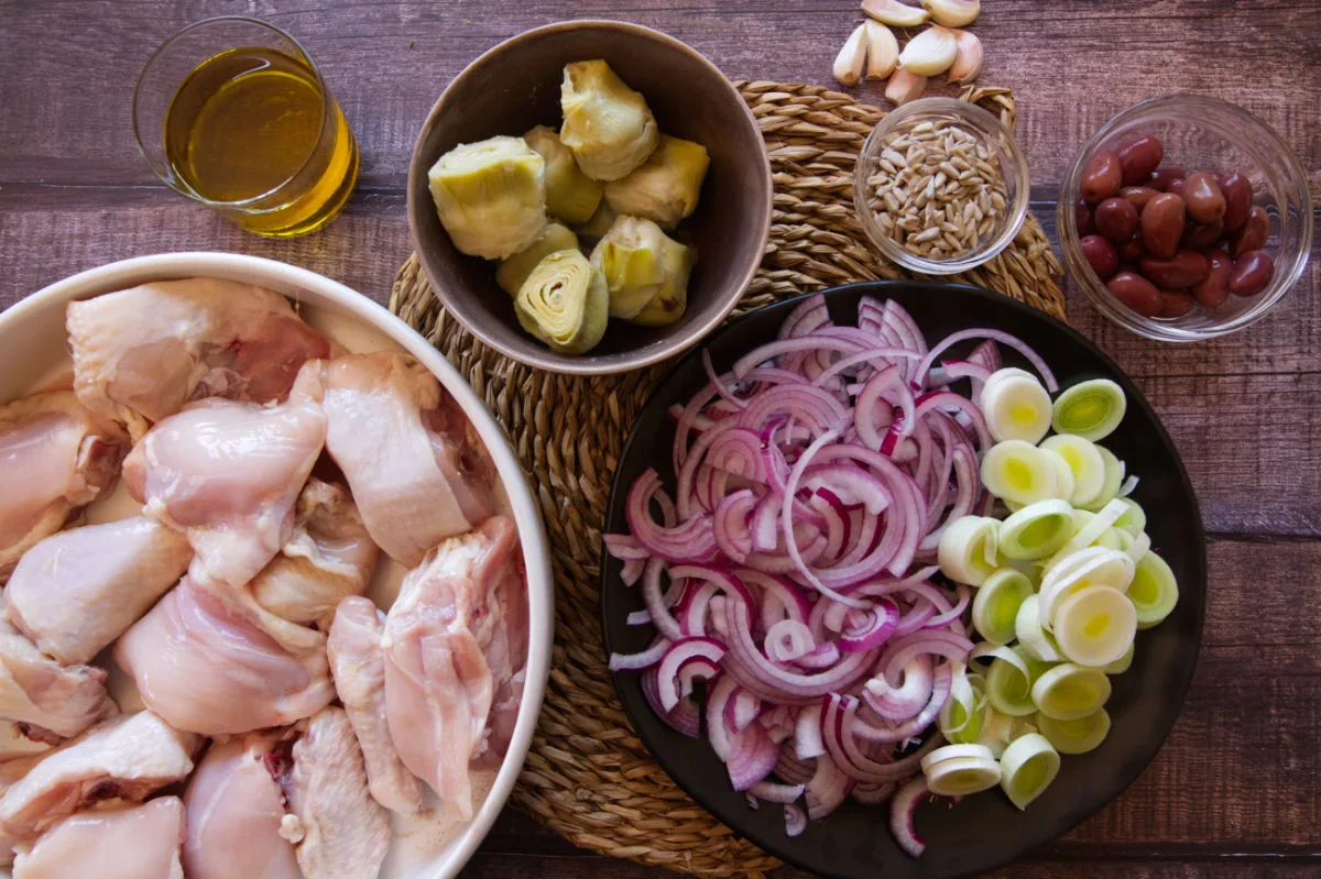 Chicken pieces, leek, red onion, and artichoke hearts sit on a kitchen counter.