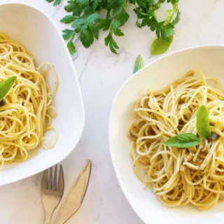 Two bowls of spaghetti with anchovies and capers sit beside fresh parsley.