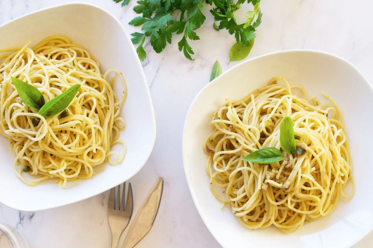 Two bowls of spaghetti with anchovies and capers sit beside fresh parsley.