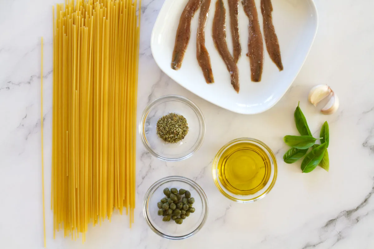 A small plate of anchovies sits beside some spaghetti, capers, olive oil, and basil.