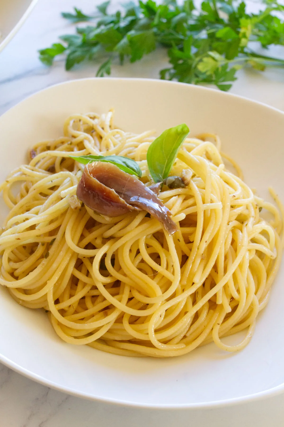 A bowl of Spaghetti with anchovies and capers is garnished with fresh basil leaves and an anchovie fillet.