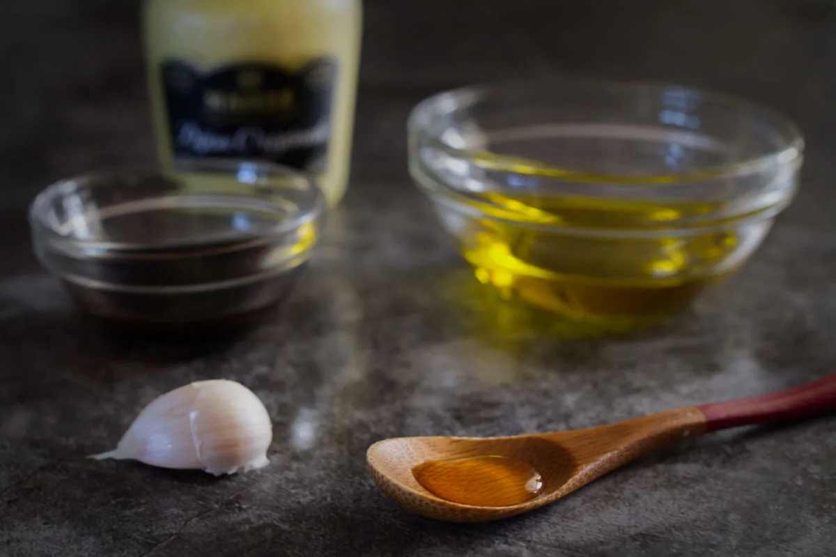 a spoonful of honey sits beside some olive oil, dijon mustard, balsamic vinegar, and a garlic clove. 