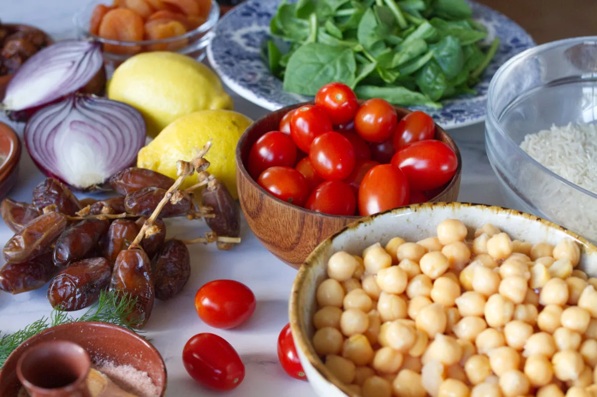 a bowl of chickpeas sits next to some dried dates, cherry tomatoes, spinach leaves, lemons, herbs, and spices.
