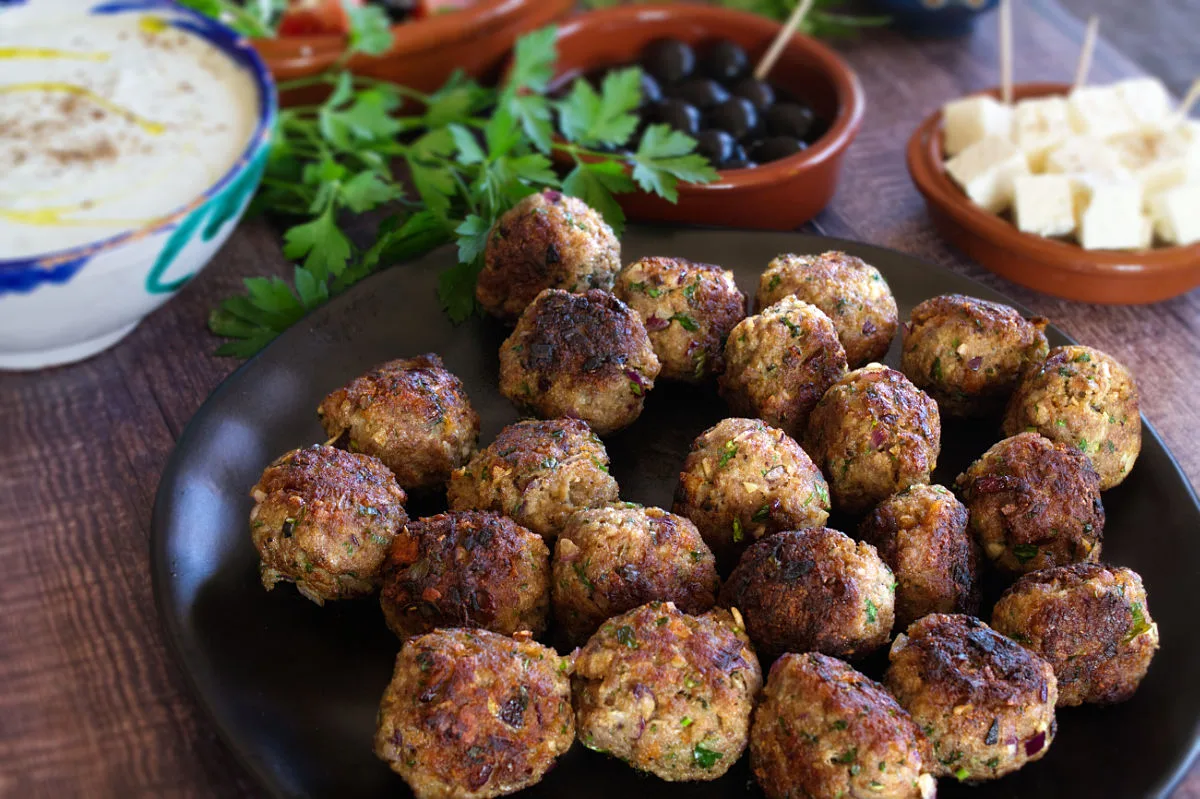 A plate of Greek meatballs sits next to some Feta cheese, Tzatziki dip, and olives.