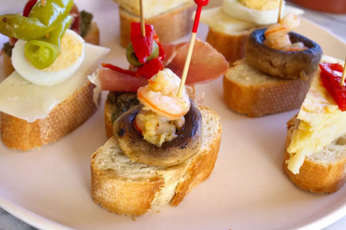 A plate with Spanish pintxos on it, the front one is bread with garlic mushrooms and a shrimp on top. 