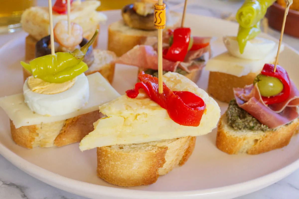 Various Spanish pintxos laid out on a plate. The pinchos in front has potato omelet and pickled red pepper on a piece of bread. 