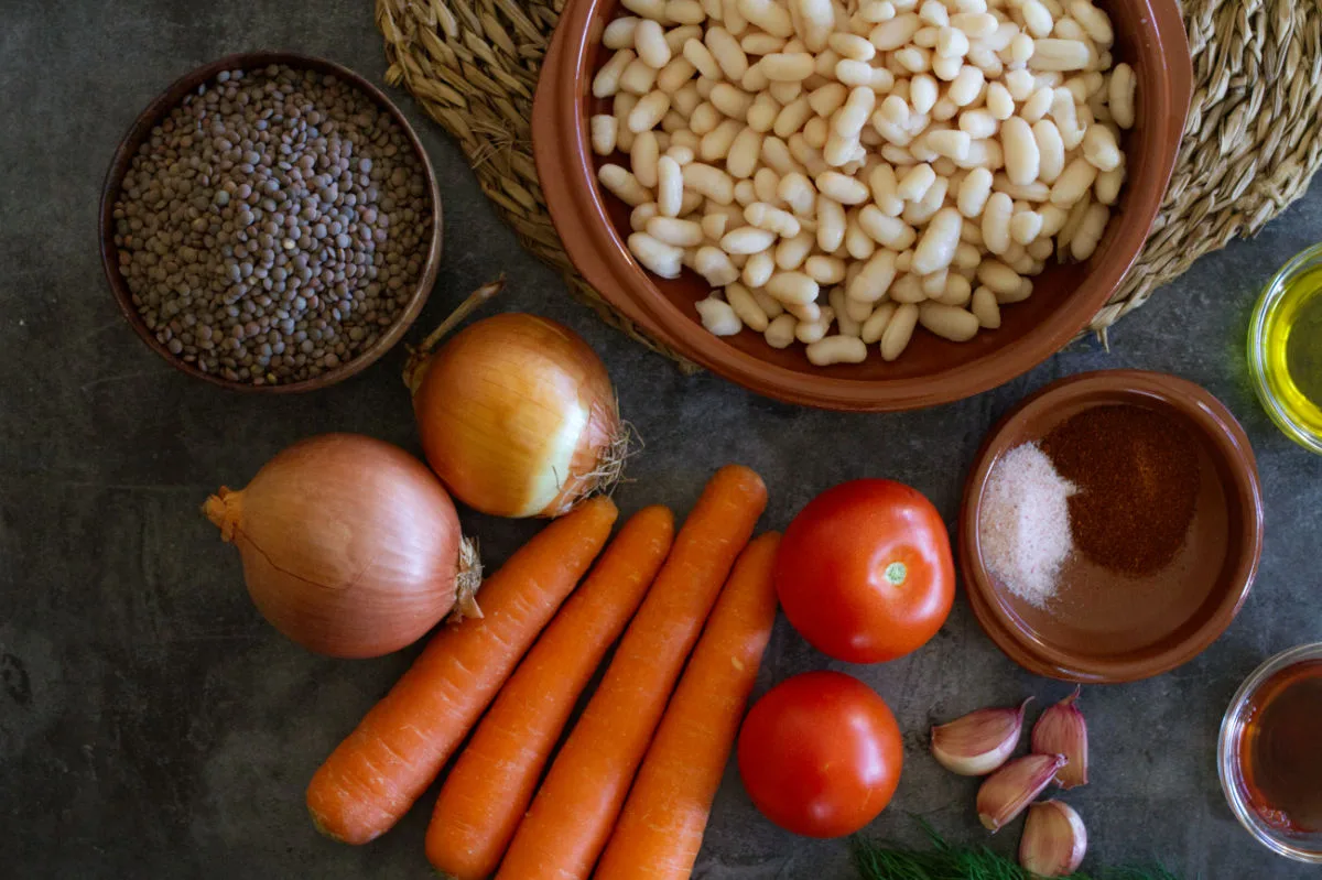 A bowl of brown lentils sits beside some vegetables, garlic, spices, and white beans.
