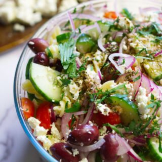 A bowl of Mediterranean pasta salad sits on a table with a garnish of feta cheese and fresh chopped parsley.