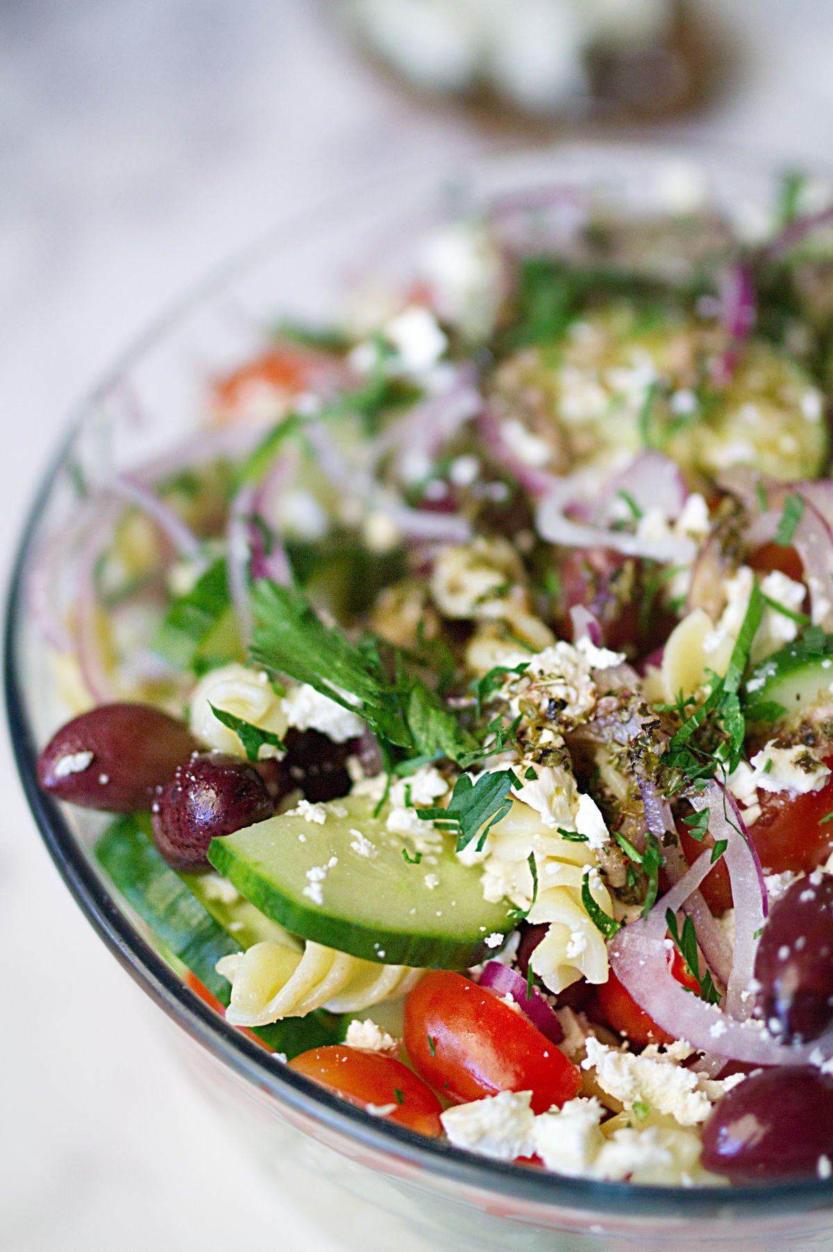 A bowl of Mediterranean pasta salad sits on a table with a garnish of feta cheese and fresh chopped parsley.