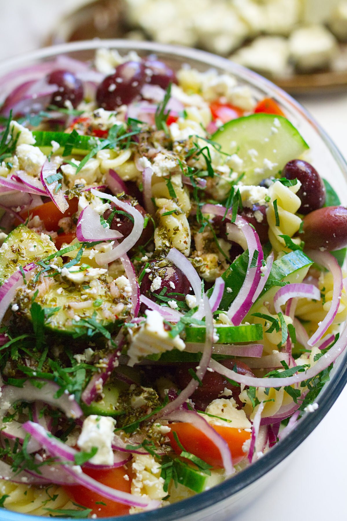 A bowl of Mediterranean pasta salad sits on a table with a garnish of feta cheese and fresh chopped parsley.