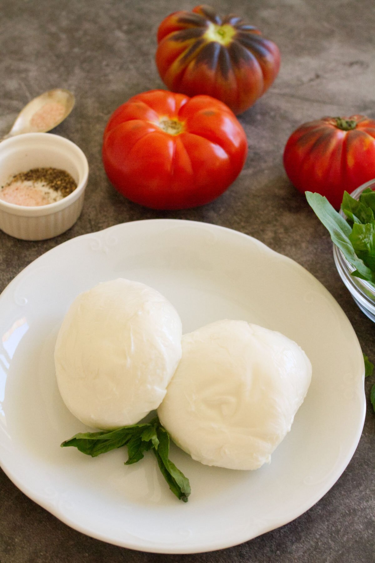 two balls of mozzarella sit beside some ripe red tomatoes and some basil.