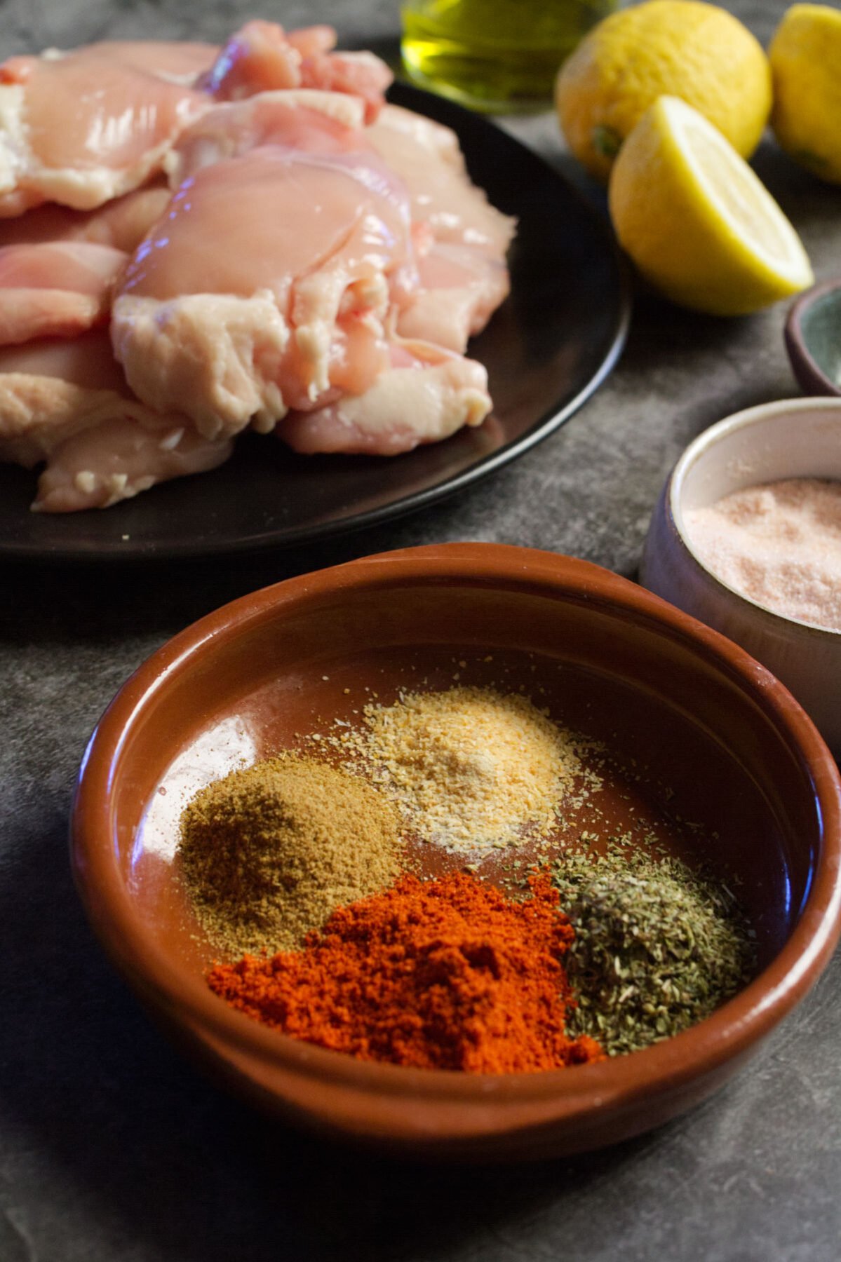 A bowl of spices sits beside some uncooked chicken thighs, lemon, and fresh parsley.