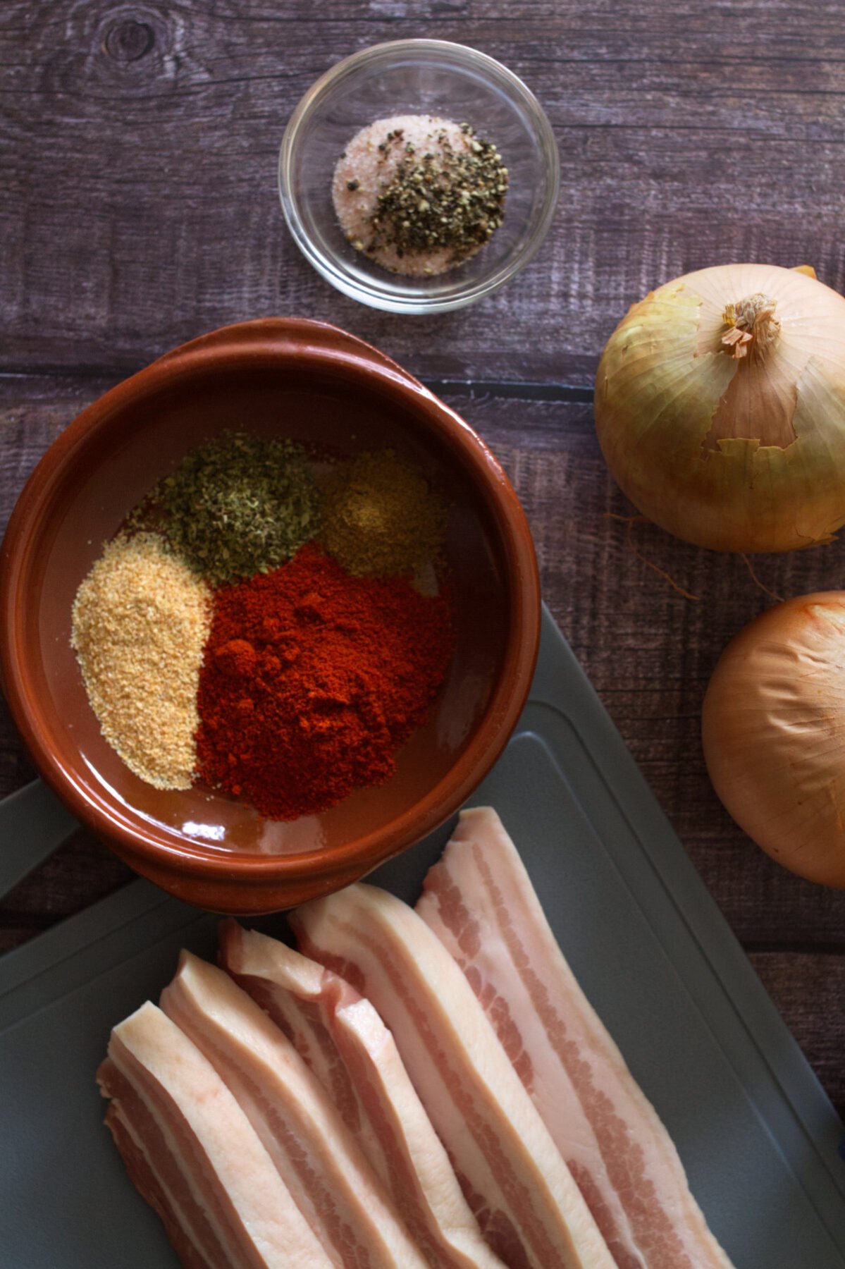 Some spices sit beside some thick cuts of pancetta and some garlic and onions.