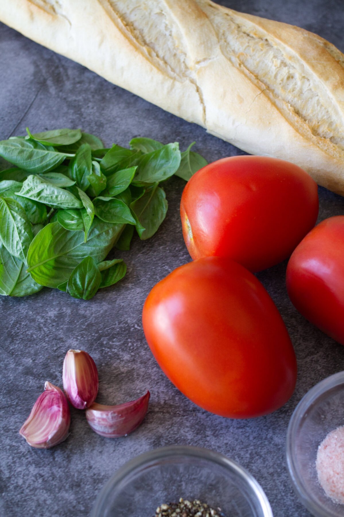 Some ripe Roma tomatoes sits beside some fresh basil leaves, garlic cloves, and bread. 