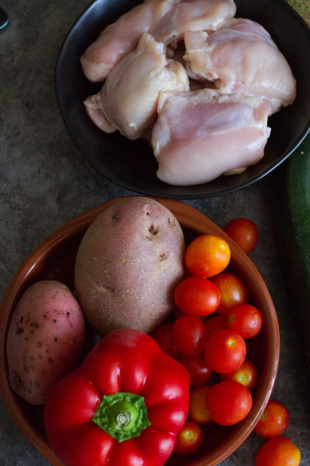 Some uncooked chicken thighs sit beside some fresh tomatoes, potatoes, and red bell pepper. 