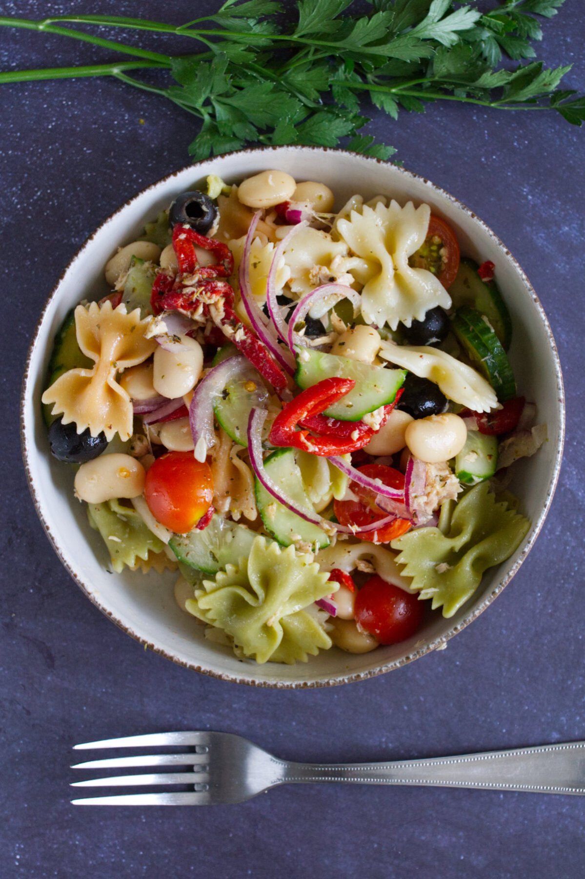 A bowl of mediterranean pasta salad with tuna sits beside a fork and some fresh parsley