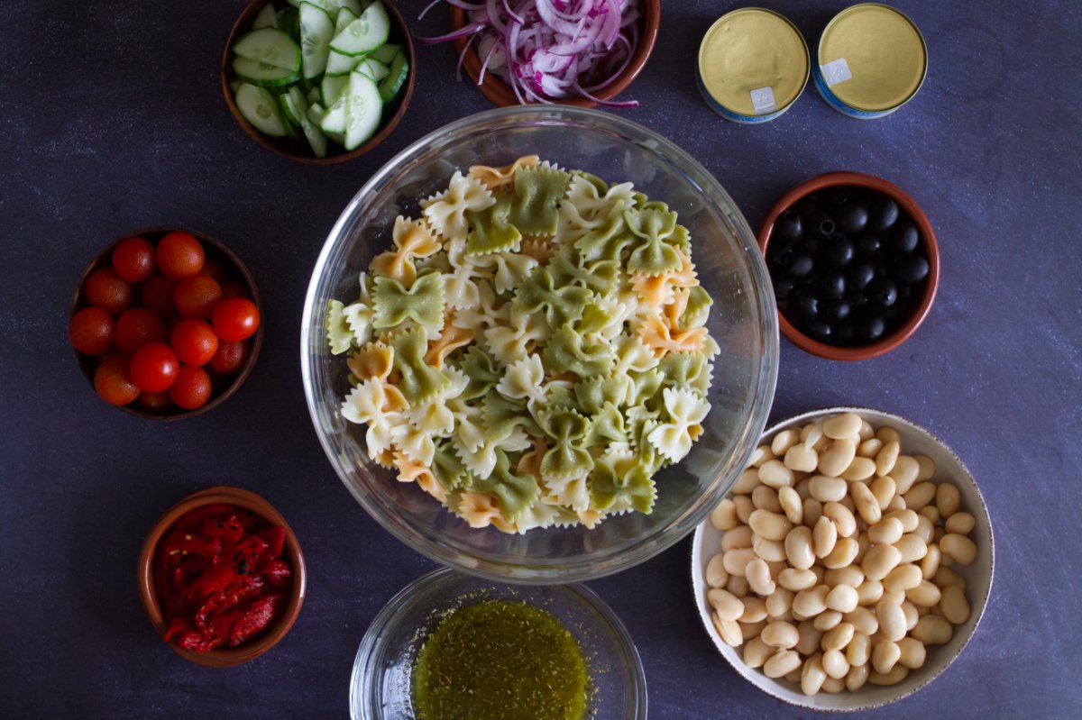 A bowl of farfalle pasta sits beside some beans, olives, veggies, and canned tuna.