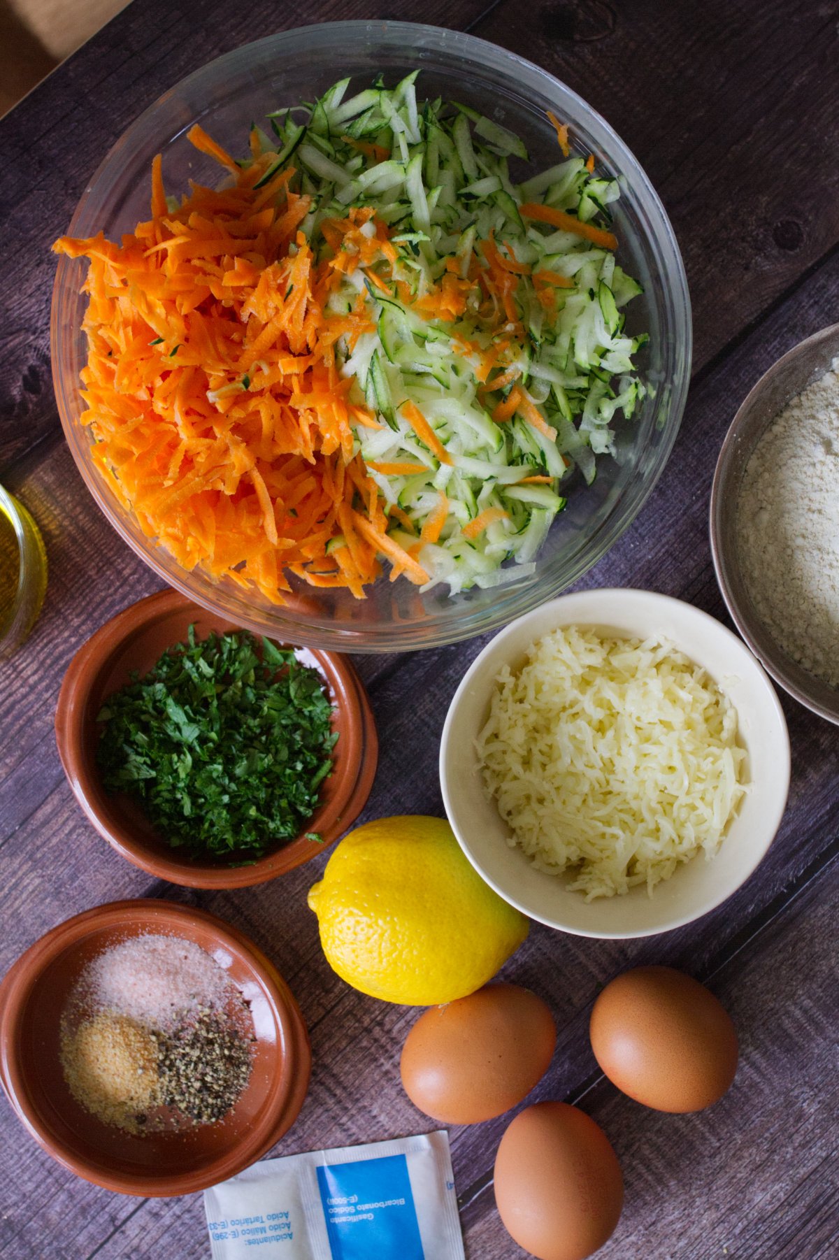 A large bowl of grated zucchini and carrot sits beside eggs, flour, mozzarella cheese, and herbs. 