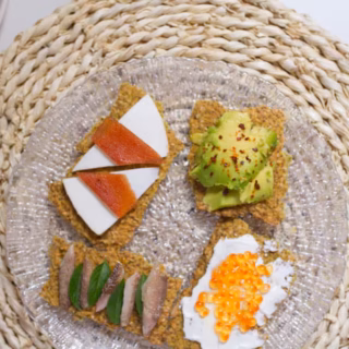 A glass plate with four homemade seed crackers on it with different toppings.